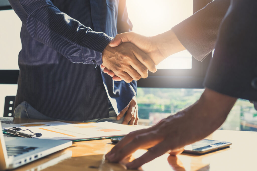 Two people shake hands over a desk with papers and a laptop, signaling agreement in a business meeting.