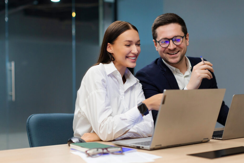 Two positive colleagues engaging in discussion, using laptop for project collaboration in modern office environment, sitting at desk in board room